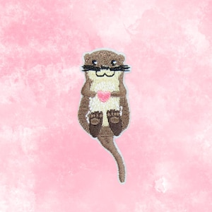 May include: Embroidered patch featuring a cartoon otter holding a pink heart. The otter has brown fur, a cream-colored belly, and black whiskers. The patch is set against a pink watercolor background.