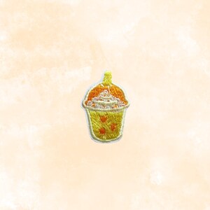 May include: Embroidered patch of a bubble tea drink. The patch features a yellow cup with orange and white details, and small red heart-shaped accents. The top of the drink is orange with white whipped cream.