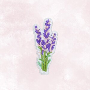 May include: Embroidered patch featuring a lavender sprig. The patch has purple flower heads with pink accents, green stems, and a white border. The background is a soft pink watercolor wash.