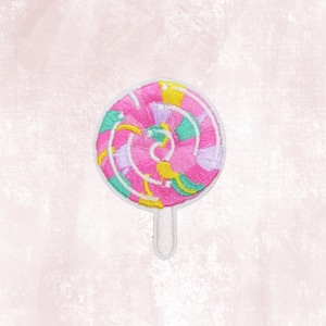 May include: Embroidered patch in the shape of a pink lollipop with a white stick. The lollipop design features a spiral pattern with shades of pink, green, yellow, and purple. The patch has a white border and is set against a soft pink background.