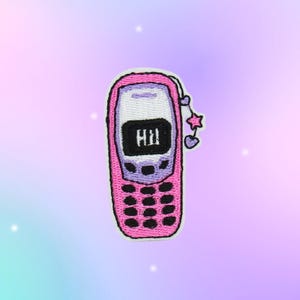 May include: An embroidered patch shaped like a retro mobile phone in shades of pink and light purple. The screen displays the text Hi in black. A small dangling charm featuring two purple hearts and a pink star is attached to the side of the phone.