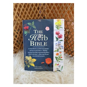 May include: A hardcover book titled "The Herb Bible" with a dark blue cover. The book features colorful illustrations of herbs and flowers. The text includes information on growing, harvesting, and preserving herbs. The book is on a fluffy white surface.
