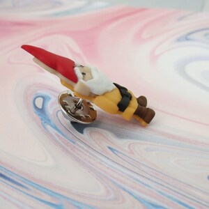 May include: A small, red-hatted gnome figurine with a white beard and yellow clothing. The gnome is wearing a black belt and brown shoes. The figurine is attached to a silver pin.