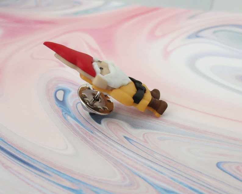 May include: A small, red-hatted gnome figurine with a white beard and yellow clothing. The gnome is wearing a black belt and brown shoes. The figurine is attached to a silver pin.