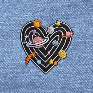 May include: A heart-shaped embroidered patch featuring a black background with white concentric heart outlines. The design includes colorful celestial bodies such as a ringed planet, a yellow sun, a shooting star, and various small planets and stars.