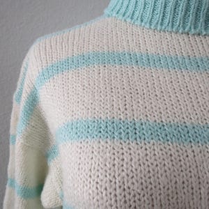 May include: A hand-knitted sweater featuring a ribbed mock neck collar and horizontal stripes in alternating cream and light turquoise blue. The garment is constructed with a chunky knit texture, showcasing a consistent stitch pattern throughout.