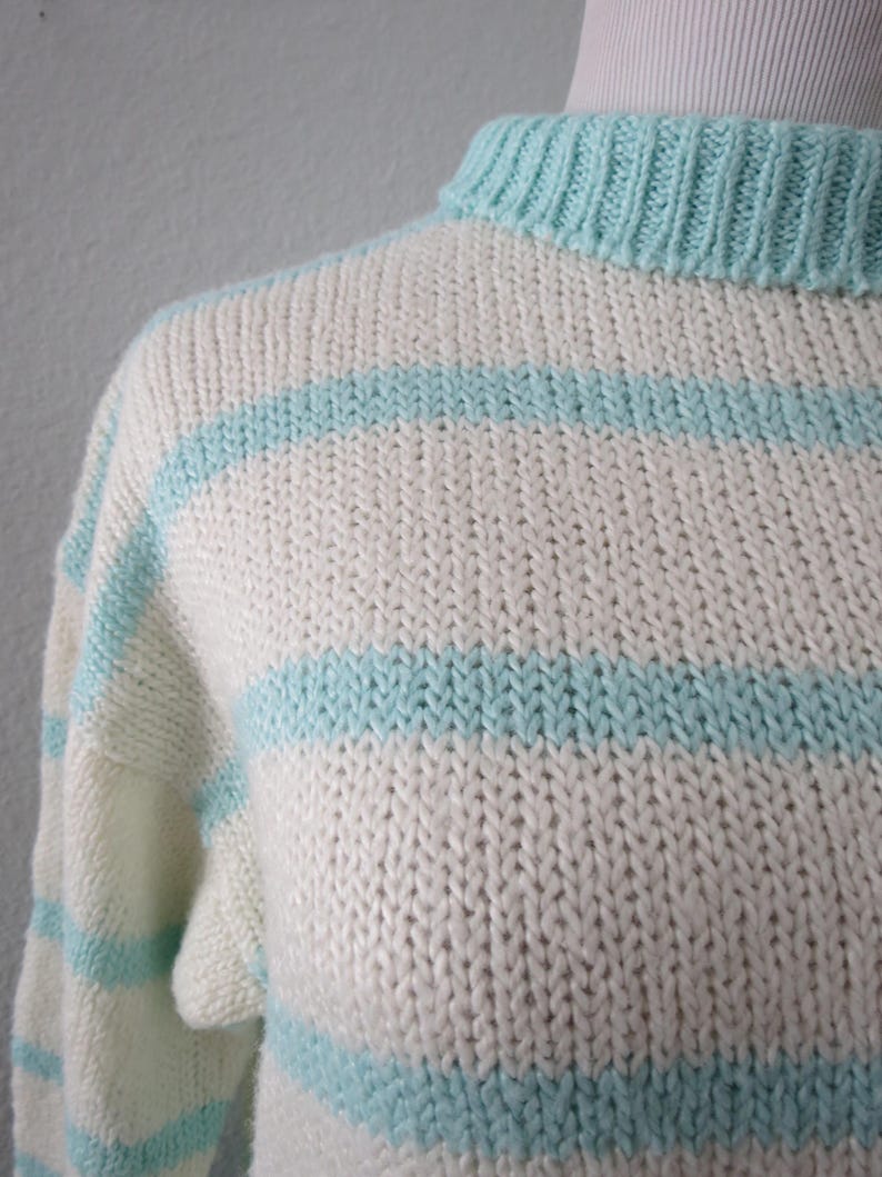 May include: A hand-knitted sweater featuring a ribbed mock neck collar and horizontal stripes in alternating cream and light turquoise blue. The garment is constructed with a chunky knit texture, showcasing a consistent stitch pattern throughout.