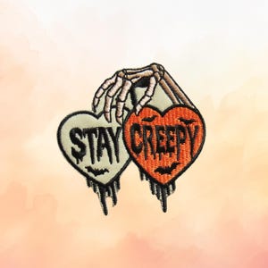 May include: An embroidered patch featuring a skeletal hand holding two heart-shaped designs. The left heart is cream-colored with the text STAY, and the right heart is orange with the text CREEPY. Both hearts feature black dripping details and small bat silhouettes.