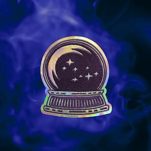 May include: A circular embroidered patch featuring a dark purple crystal ball design with a crescent moon and several small four-pointed stars inside. The patch has a reflective, iridescent border that catches light with rainbow hues.