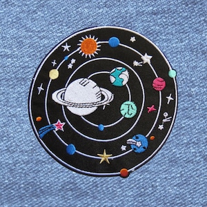 May include: A circular embroidered patch featuring a black background with a white solar system design. The patch displays a large white planet with rings, various colorful planets in shades of blue, green, pink, and orange, a bright yellow sun, and small white stars scattered throughout the orbital paths.