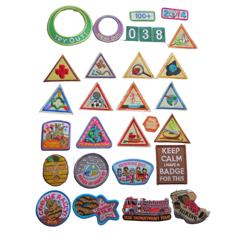 May include: A collection of embroidered patches in various shapes and colors. Includes patches with text like "TRY OUT!", "COOKIES", and "HIKING". The patches feature images of a fire truck, a beehive, and a hiking boot.