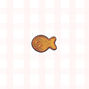 May include: Embroidered patch in the shape of a smiling goldfish. The patch is primarily gold with a black outline and a small, smiling face. The background is a light pink and white checkered pattern.