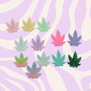 May include: A collection of colorful, cannabis leaf-shaped embellishments. The leaves are in various pastel shades, including pink, yellow, green, and purple, with some featuring glitter. The background has a wavy, lavender and white pattern.