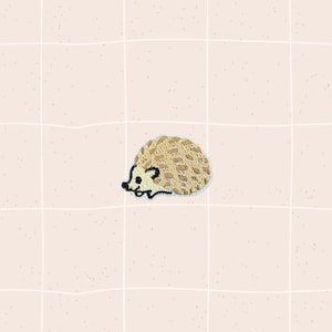 May include: A small, embroidered patch featuring a cartoon hedgehog. The hedgehog has a tan body with a textured pattern and a black and white face. The patch is likely designed to be ironed or sewn onto fabric.