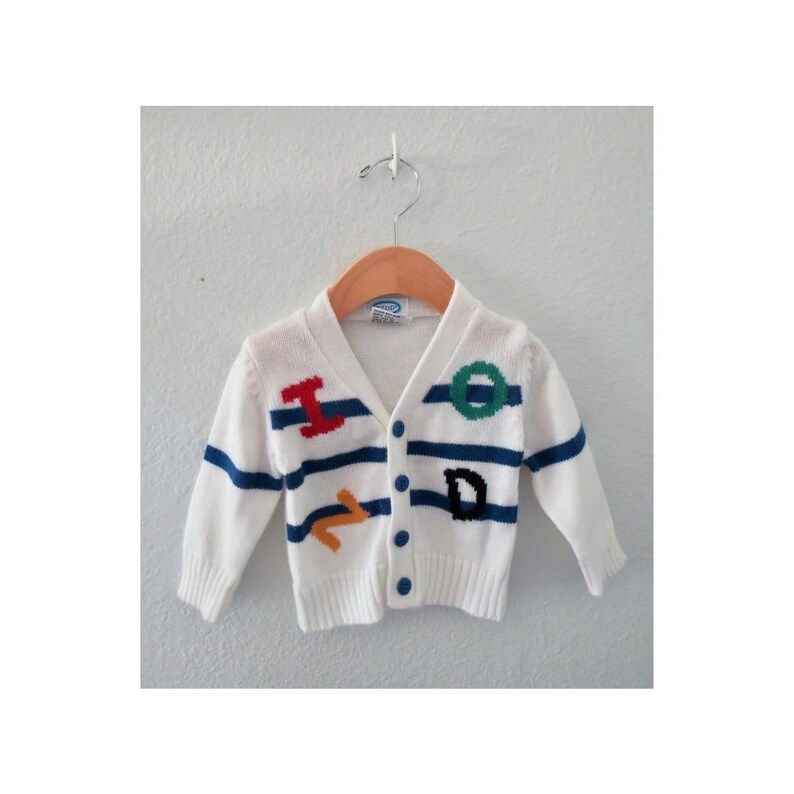 May include: A white long-sleeve knit cardigan featuring horizontal blue stripes and embroidered letters in red, green, and orange. The garment has a V-neckline, a front button closure with four blue buttons, and ribbed cuffs and hem on a hanger.