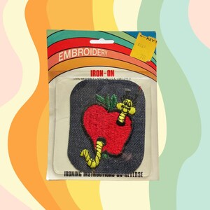 May include: Iron-on patch featuring a red apple with a green stem and a yellow worm with a black face eating its way into the apple. The patch is on a blue denim background. The packaging says "Embroidery Iron-On 100% Washable Made in USA Valdyl".