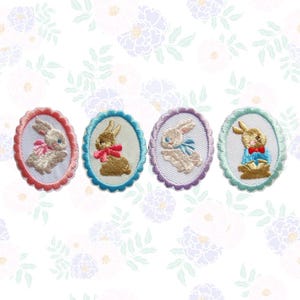 May include: Four embroidered oval patches featuring Easter bunnies. Each patch has a different color border: pink, blue, purple, and green. The bunnies are in various colors with bows and outfits. The background is white with a floral pattern.