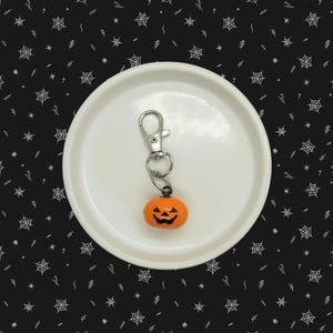 May include: A silver-toned metal lobster clasp keychain featuring a small, round, orange pumpkin charm. The pumpkin has a black, pixelated jack-o'-lantern face with triangular eyes and a jagged mouth, suspended from a circular metal jump ring.