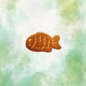May include: Embroidered patch in the shape of a fish, in shades of orange and brown. The fish has detailed stitching, including scales and fins. The background is a watercolor wash of white, green, and yellow.