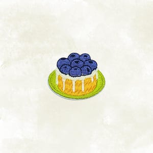 May include: Embroidered patch featuring a miniature cake topped with blueberries. The cake has a yellow base with white frosting drips and sits on a green plate. The blueberries are a deep blue color, and the patch has a stitched texture.