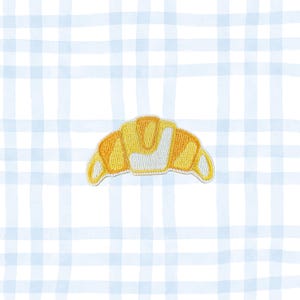 May include: A yellow and white embroidered croissant patch. The patch is in the shape of a crescent roll with a detailed design. The background is a light blue and white checkered pattern.