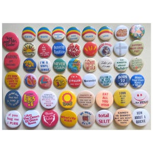 May include: A collection of circular pin-back buttons arranged in rows, featuring various colors and printed text slogans such as 'Cats', 'Tofu', 'Peace', 'Love', 'Born to Shop', 'Total Slut', and 'How About a Quickie' alongside graphic illustrations.