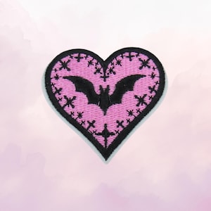May include: A heart-shaped embroidered patch featuring a pink background with a black border. The center displays a black bat silhouette surrounded by small black cross symbols arranged in a pattern across the pink surface of the heart.