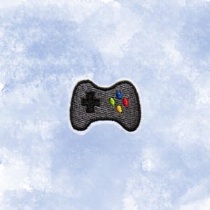 May include: Embroidered patch of a black video game controller with a white border. The controller features a black cross, and colorful buttons in red, blue, yellow, and green. The patch is set against a light blue background.