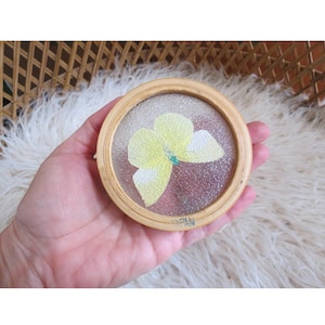Puede incluir: Posavasos redondo con marco de madera clara y centro de vidrio texturizado. El vidrio presenta un dise&ntilde;o de flor amarilla con detalles blancos y un centro azul. El posavasos est&aacute; marcado "JAPAN".