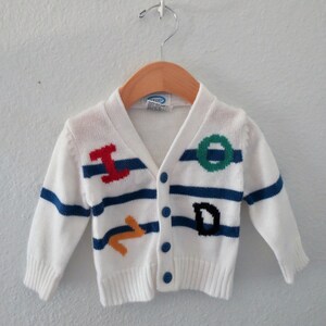 May include: A white long-sleeve knit cardigan featuring horizontal blue stripes and colorful embroidered letters. The front includes a red I, a green O, a yellow Z, and a black D. The garment has a V-neckline, ribbed cuffs, and four blue buttons.