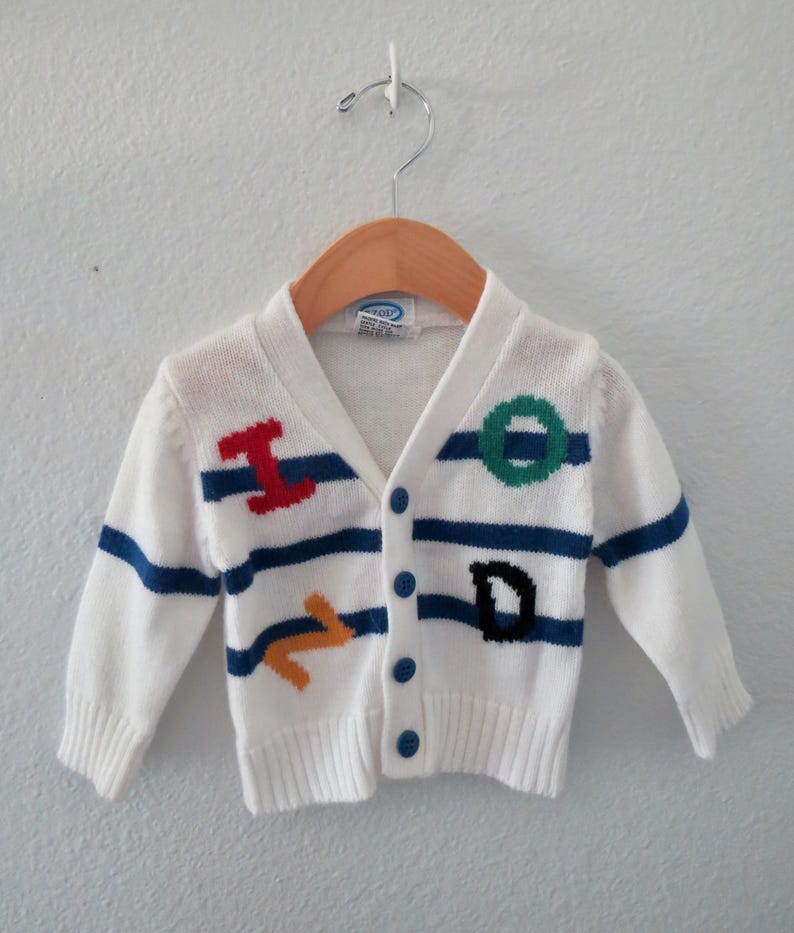 May include: A white long-sleeve knit cardigan featuring horizontal blue stripes and colorful embroidered letters. The front includes a red I, a green O, a yellow Z, and a black D. The garment has a V-neckline, ribbed cuffs, and four blue buttons.