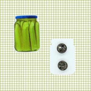 May include: A jar of pickles with a blue lid and a white enamel pin with two pin backs. The pickles are bright green and the jar is clear. The pin is shaped like a jar of pickles.