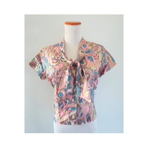 May include: A short-sleeved blouse featuring a vibrant paisley pattern in shades of pink, blue, and yellow. The garment includes a V-neckline with an integrated tie detail at the center front, creating a soft drape. The fabric has a smooth finish.