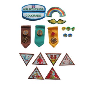 May include: A collection of Girl Scout patches and pins. Includes patches with text "Girl Scouts USA" and "Colorado", a rainbow patch, and various triangular patches with animal designs. Also, there are several pins and ribbon-like badges.