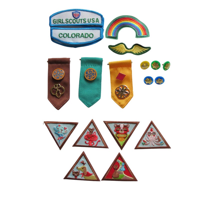 May include: A collection of Girl Scout patches and pins. Includes patches with text "Girl Scouts USA" and "Colorado", a rainbow patch, and various triangular patches with animal designs. Also, there are several pins and ribbon-like badges.