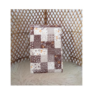 May include: A rectangular patchwork quilt with a mix of brown, white, and floral patterned squares. The quilt is resting on a fluffy, white surface, with a woven, diamond-patterned backdrop.