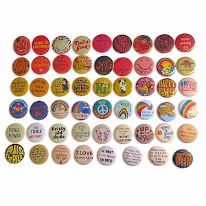 May include: A collection of circular pin-back buttons arranged in rows, featuring various colorful graphic designs, illustrations, and text slogans including phrases like 'Feelin Good', 'Praise the Pill', 'Pickle Power', and 'Smile'.
