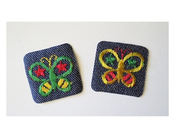 May include: Two embroidered butterfly patches. Each patch is a dark blue square with a butterfly design. One butterfly is green, yellow, and red. The other is yellow, green, and red. The patches are on a white background.