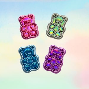 May include: Four embroidered patches shaped like gummy bears, each featuring a metallic, iridescent finish. The patches are colored in vibrant shades of pink, green, blue, and purple, with stitched borders outlining the bear-shaped silhouettes.