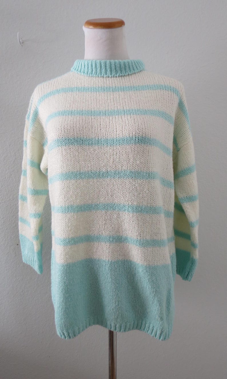 May include: A long-sleeve knit sweater featuring a ribbed mock neck collar. The garment displays a horizontal striped pattern in alternating bands of off-white and light turquoise blue, with a solid light turquoise blue hem at the bottom.