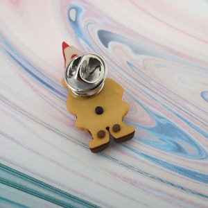 May include: A brown and yellow gnome pin with a red hat. The pin has a silver back with a safety clasp.