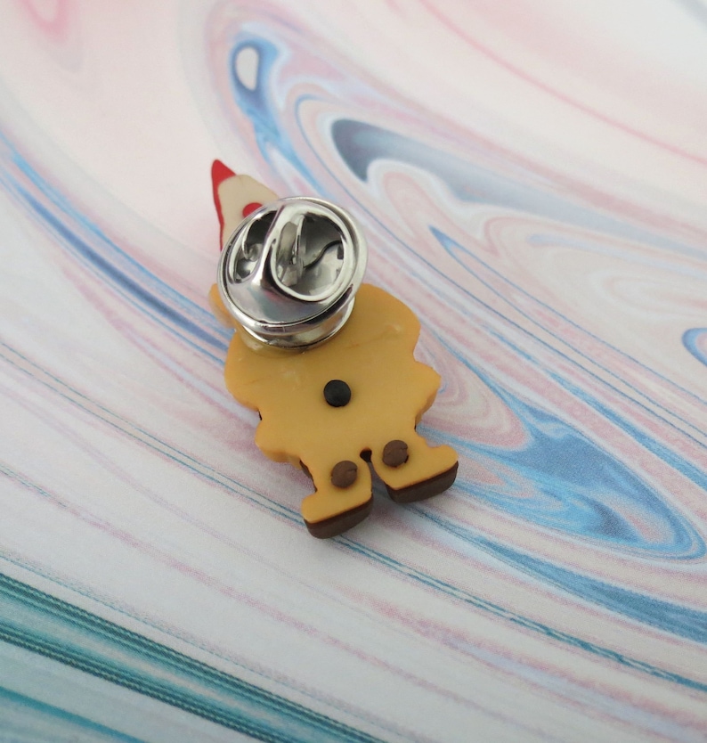 May include: A brown and yellow gnome pin with a red hat. The pin has a silver back with a safety clasp.