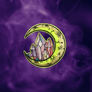 May include: An embroidered patch featuring a bright yellow crescent moon with black line art details. Nestled within the curve of the moon is a cluster of multi-faceted crystals in shades of pink, purple, white, and teal with black outlines.