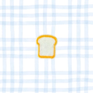 May include: A white bread slice patch with a yellow border, centered on a light blue and white plaid background. The patch has a textured appearance, suggesting an embroidered or felt design. The overall aesthetic is simple and cute.