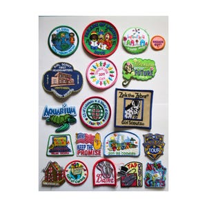 May include: A collection of colorful embroidered patches with various designs. Patches include text like "Girl Scout," "Aquarium," "Parade of Lights," and "Keep the Promise." The patches feature images of a zebra, a turtle, and a gingerbread house.