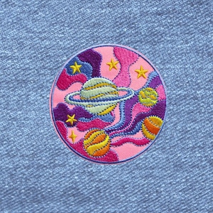May include: A circular embroidered patch featuring a space theme with a central ringed planet, two smaller spherical planets, and several yellow stars. The background consists of swirling patterns in shades of pink, purple, and blue.