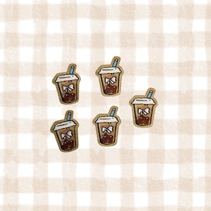 May include: Five embroidered patches in the shape of bubble tea cups. Each patch features a brown drink, a white lid, a blue straw, and a pink bow. The patches are arranged on a beige and white checkered background.