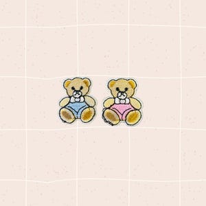 May include: Two embroidered teddy bear patches. One bear wears blue overalls and the other wears pink overalls. Both have a white bow tie and are outlined in gold thread. The background is a light pink grid.