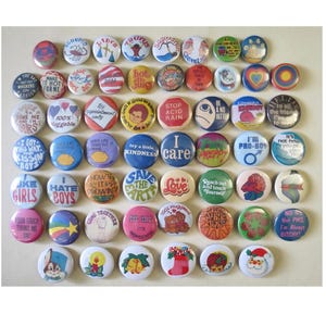 May include: A collection of circular pin-back buttons featuring various colorful graphic designs, illustrations, and text slogans including phrases like 'I care', 'Save the Earth', 'Love', 'I'm Pro-Boy', and 'Not PMS, I'm Always Bitchy'.