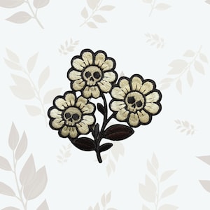May include: An embroidered patch featuring three cream-colored flowers with dark brown outlines and centers shaped like human skulls. The design includes dark brown stems and leaves, with the entire floral arrangement stitched in a cohesive cluster.